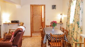 A dining area with a checkered tablecloth, wooden chairs, a vase with flowers, a clock on the wall, and a door in the background. - At the Crossroads (Louisburgh)