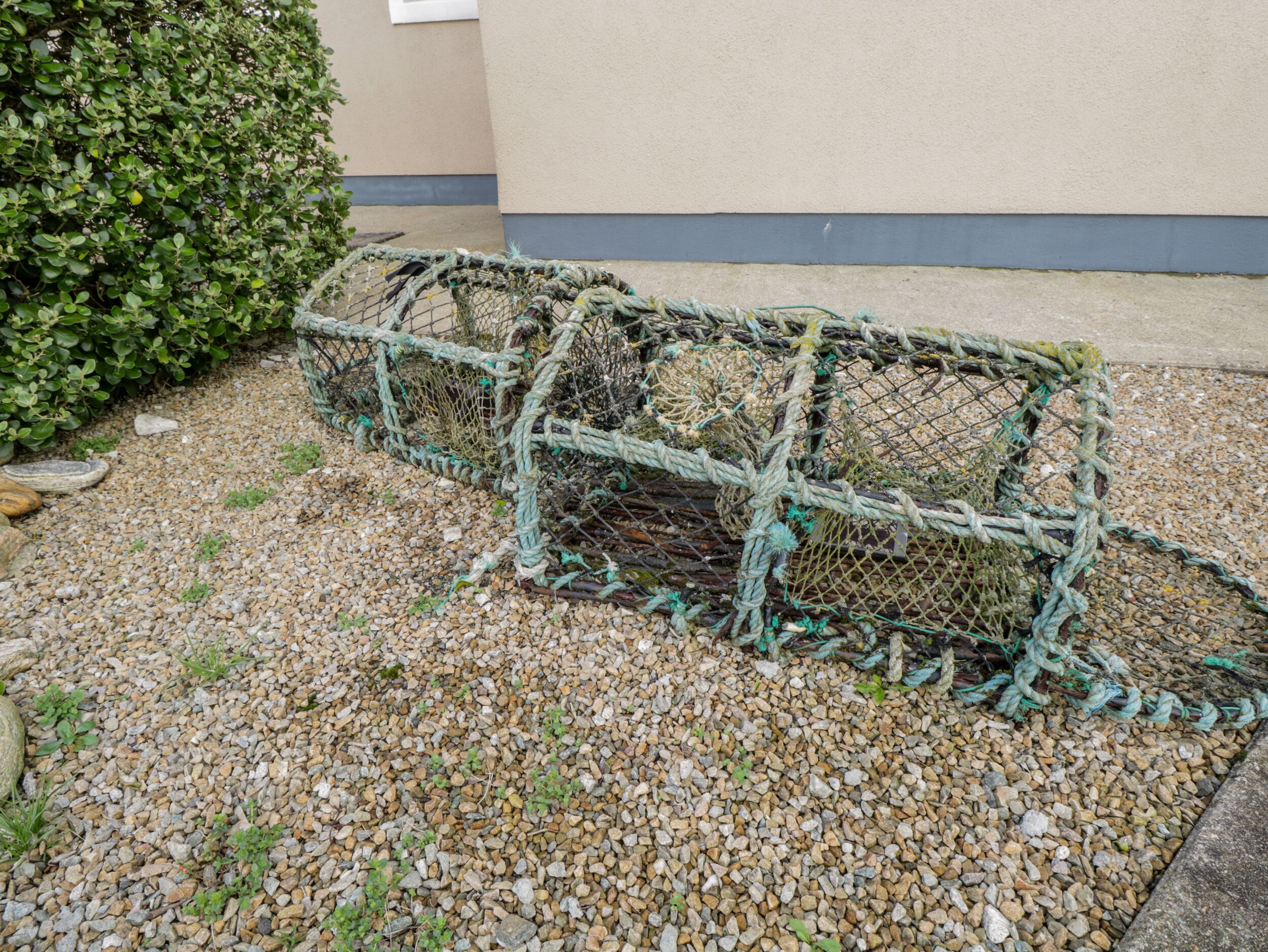 A fishing trap made of netting and rope, placed on a gravel surface near a building.