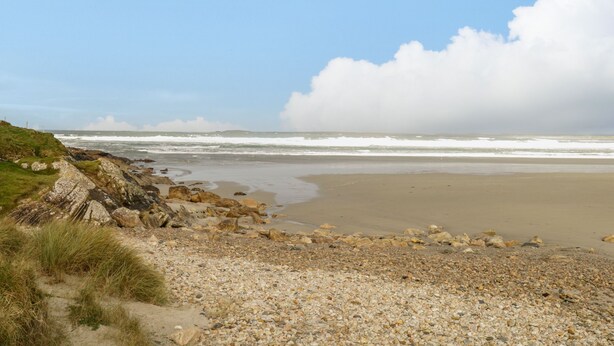A coastal beach with pebbles and grass, a rocky cliff, and waves in the background.