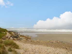 Een kuststrand met kiezels en gras, een rotsachtige klif en golven op de achtergrond.
