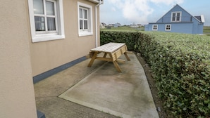 A beige house with a wooden bench outside, a hedge, and a blue house in the background.