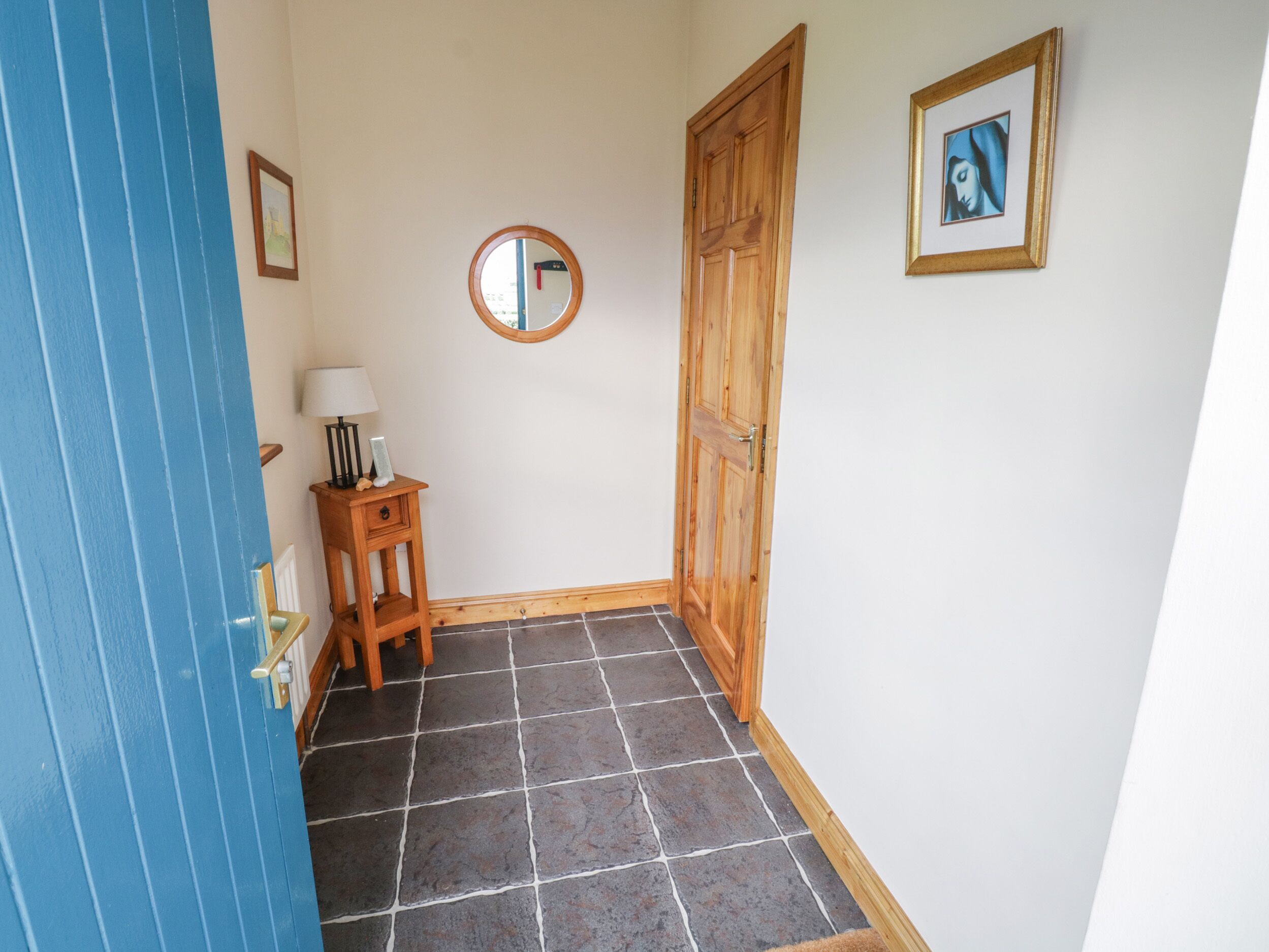 Un pasillo con una puerta azul, una puerta de madera, una mesita con una lámpara, un cuadro enmarcado y un espejo redondo en la pared.