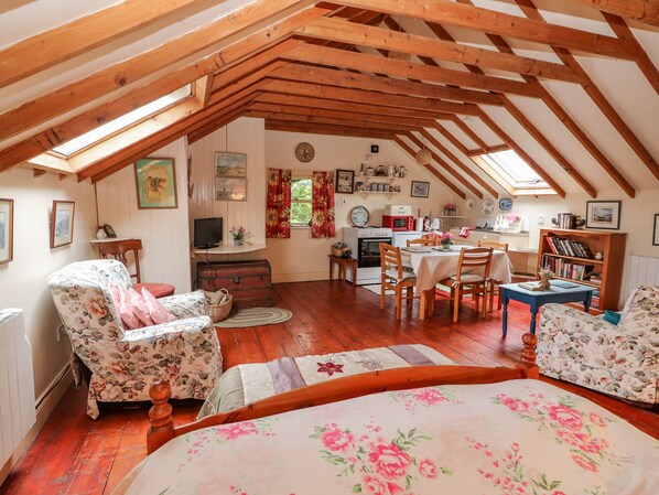 A cozy attic room with wooden beams, a dining table, chairs, a bed with floral bedding, a television, and various kitchen appliances.