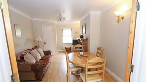 A dining area with a wooden table and chairs, a sofa with cushions, a lamp, and a window with blinds. - Aisling (Foxford)