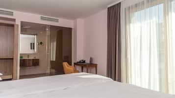 Habitación de hotel con una cama grande, un escritorio con una computadora portátil, una silla y un baño visible a través de una puerta de cristal.
