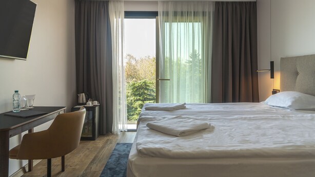 Habitación de hotel con cama, escritorio, silla, mesita de noche y vistas a árboles por la ventana.