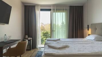 A hotel room with a bed, a desk, a chair, a nightstand, and a view of trees through the window.