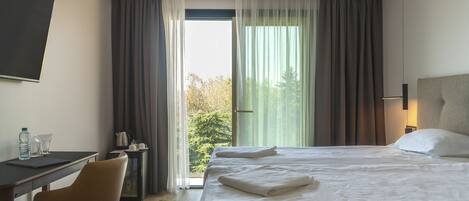 A hotel room with a bed, a desk, a chair, a nightstand, and a view of trees through the window.