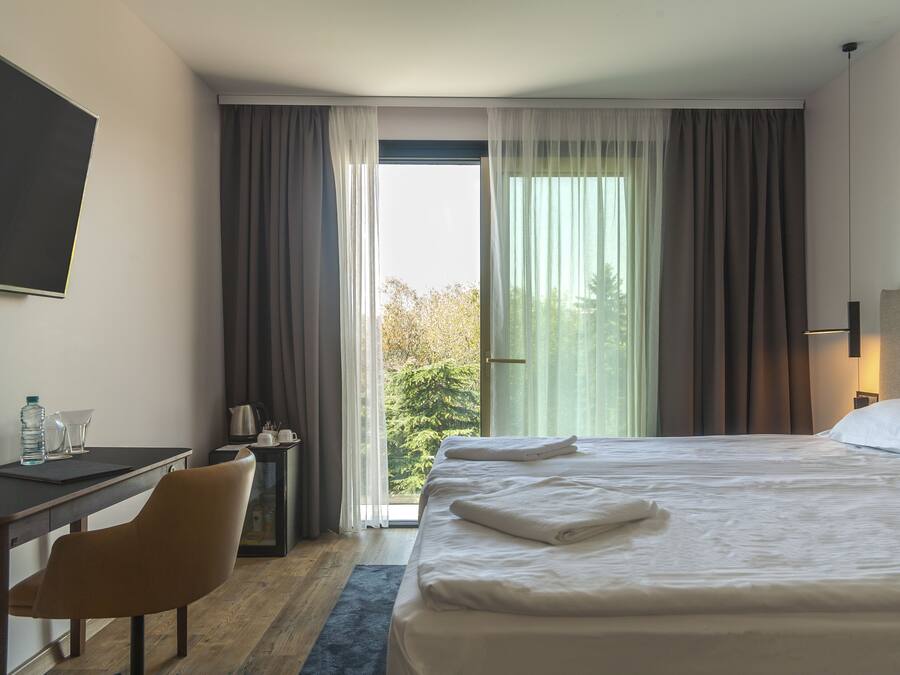 A hotel room with a bed, a desk, a chair, a nightstand, and a view of trees through the window.