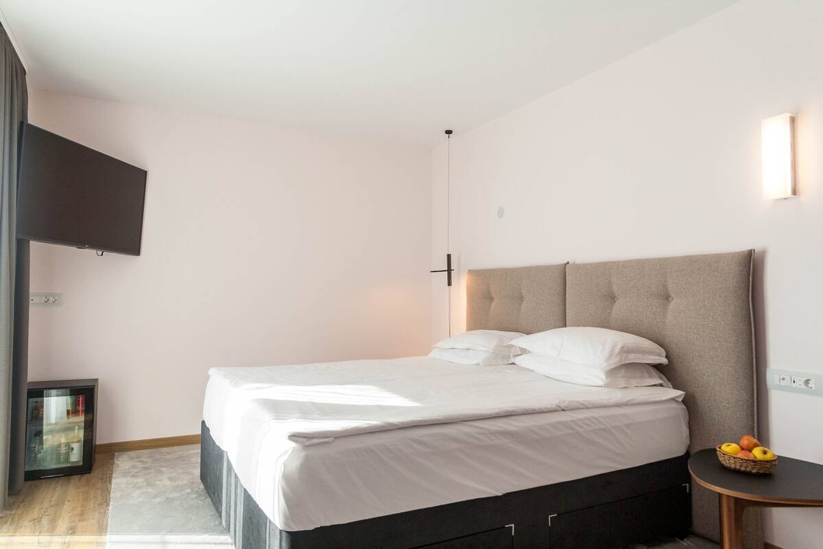 Ein ordentlich bezogenes Bett mit weißen Leinentüchern, ein kleiner Nachttisch mit Obstschale und ein an der Wand befestigter Fernseher.