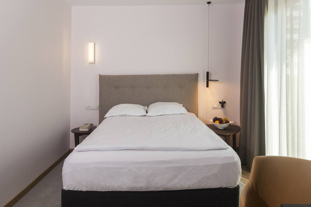 Ein Hotelzimmer mit einem großen Bett, einem Nachttisch mit Lampe, einem kleinen Tisch mit Obstschale und einem Sessel.