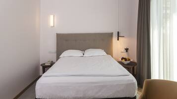 Quarto de hotel com uma cama grande, mesa de cabeceira com abajur, uma cadeira e vista para o exterior.