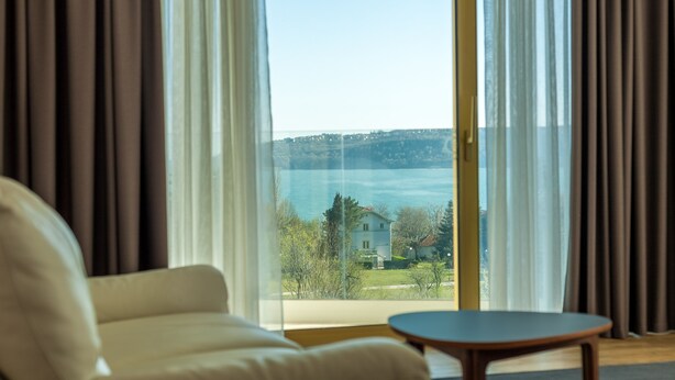 Una habitación con sofá y mesa, un ventanal con cortinas, y vistas al agua y edificios.