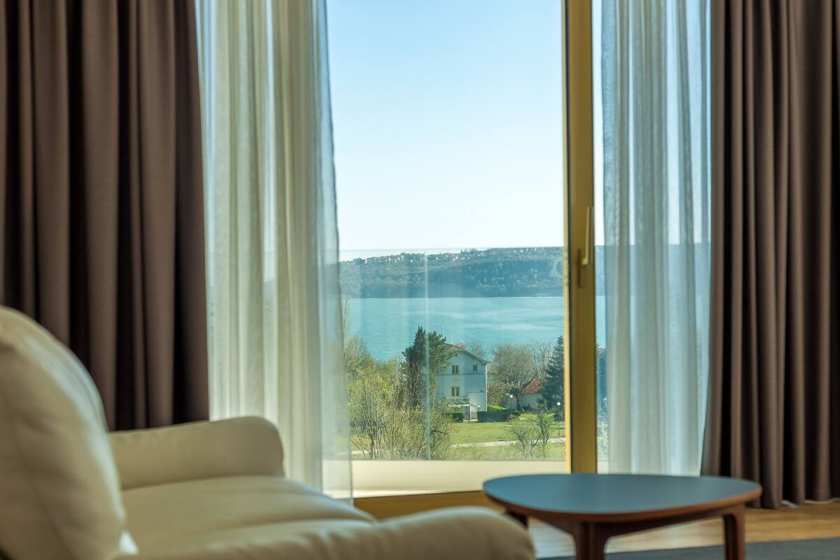 Ein Zimmer mit einer Couch und einem Tisch, ein großes Fenster mit Vorhängen und Blick auf ein Gewässer sowie Gebäude.