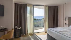 Habitación de hotel con cama, escritorio, silla, mueble pequeño y puerta de vidrio corredera que da a un balcón con vistas a árboles y una masa de agua.