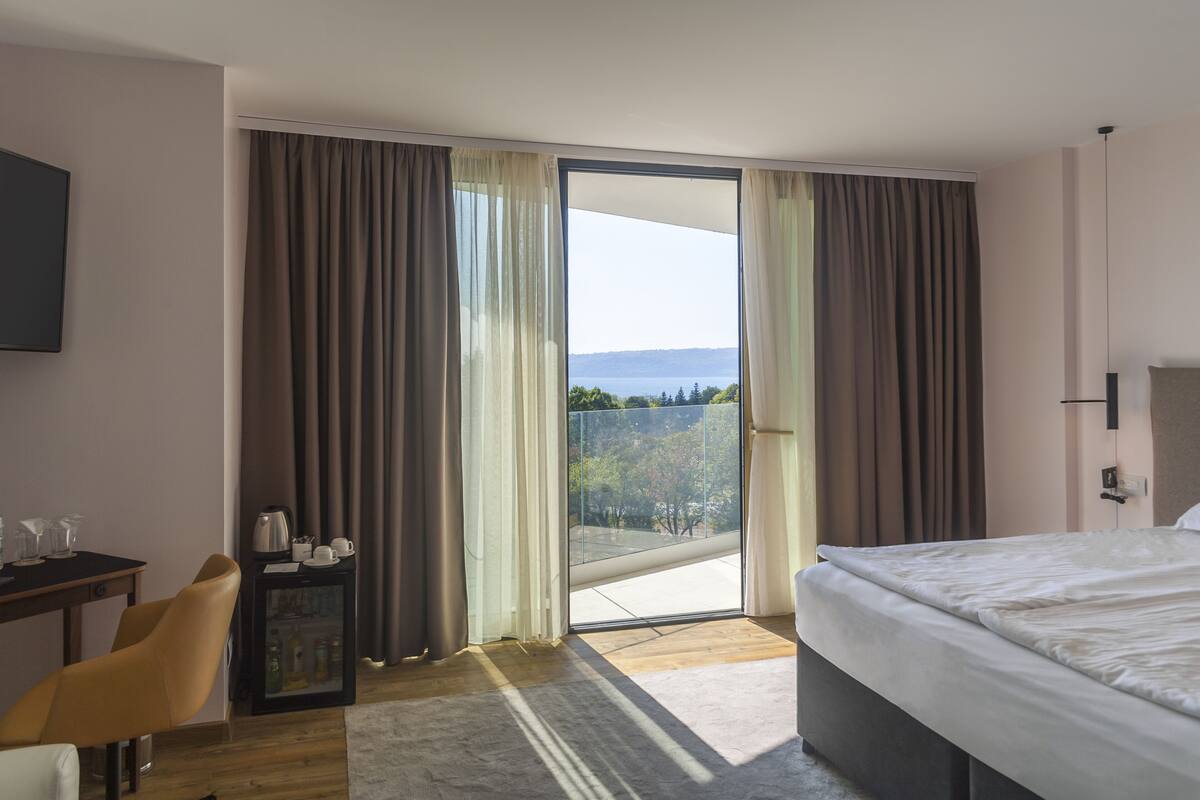 Ein modernes Hotelzimmer mit einem großen Bett, einem Schreibtisch mit Stuhl, einer kleinen Kommode und Blick ins Freie durch eine Schiebetür.
