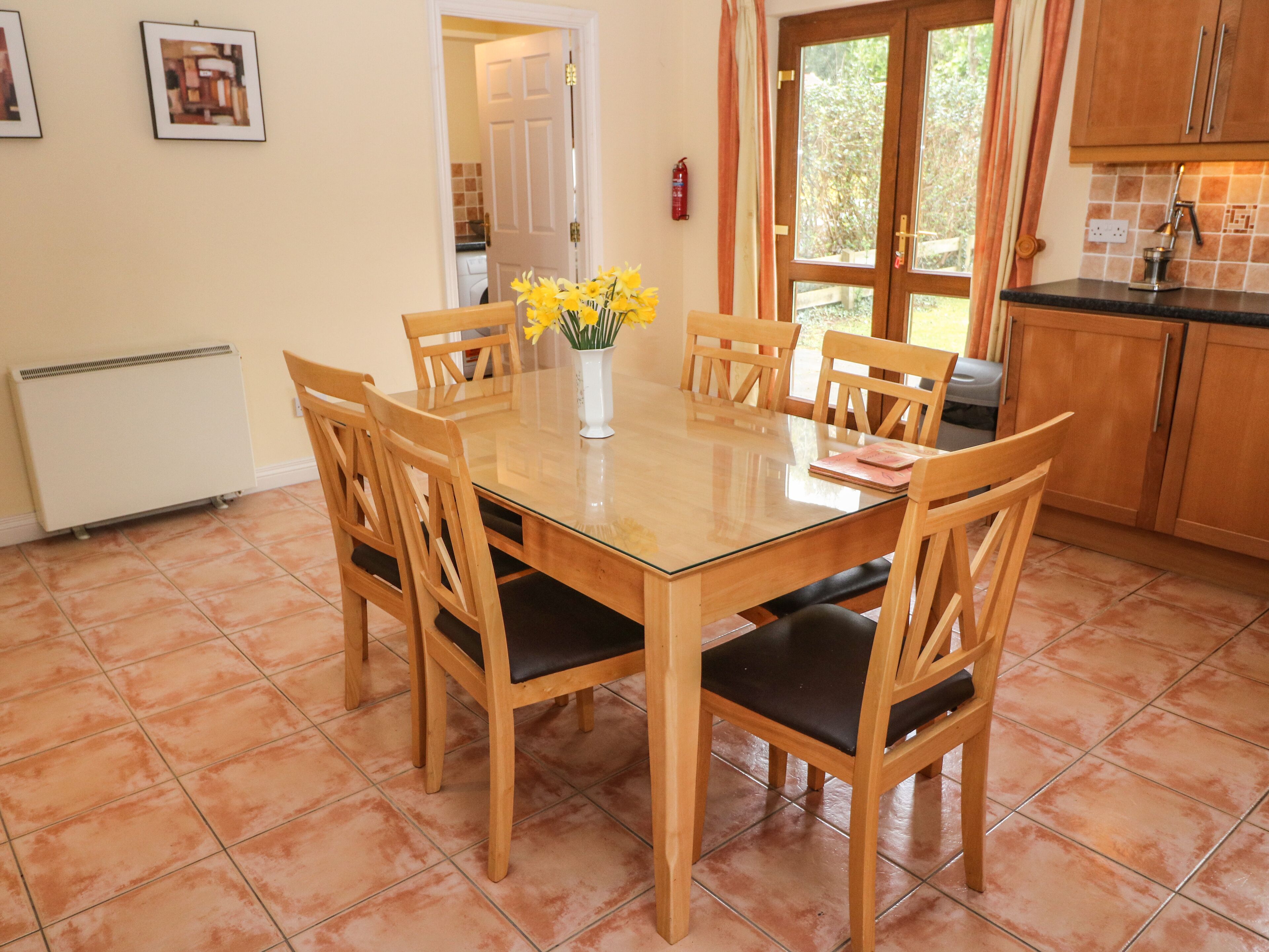 A dining area with a wooden table and chairs, a vase with flowers, and a kitchen in the background.