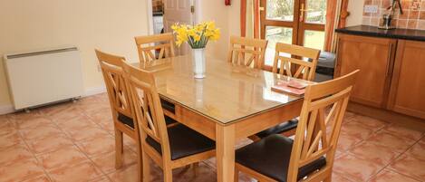 A dining area with a wooden table and chairs, a vase with flowers, and a kitchen in the background.