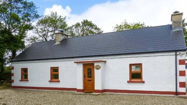 A single-story house with a dark roof, white walls, and red trim around the windows and door. - Ferrys (Portsalon)