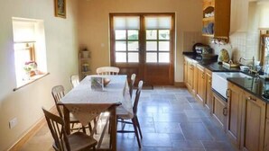 A dining area with a table and chairs, a window with blinds, and a kitchen with wooden cabinets and a countertop. - Annie's Cottage (Clogher)