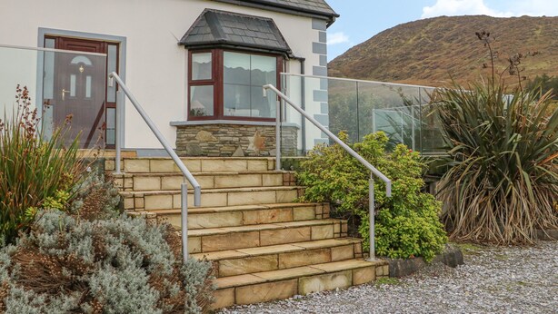 A house with a stone staircase leading to a front door, surrounded by various plants and a view of hills in the background. - Starbay House (Eyeries)