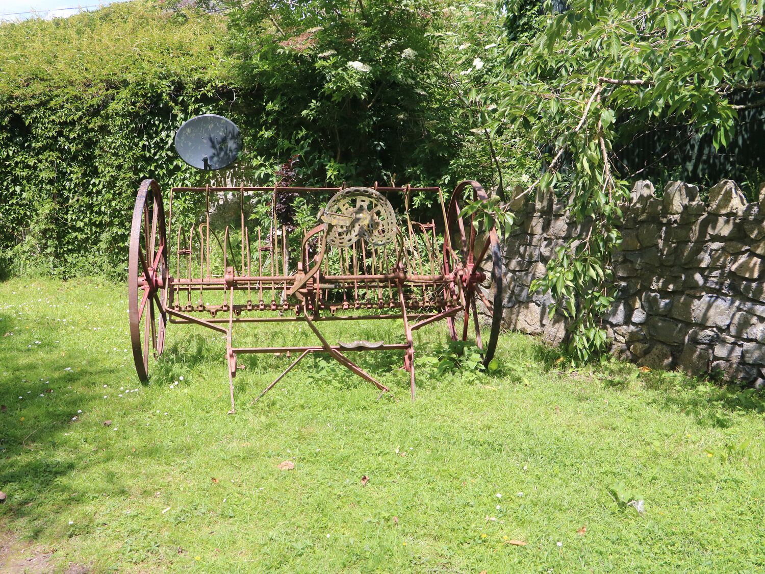 An old, rusted metal structure with wheels, possibly a historical artifact, in a grassy area with a stone wall and dense foliage in the background.