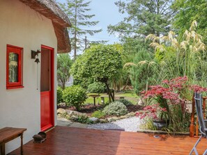 Um deck de madeira com um banco, uma porta vermelha e um jardim com diversas plantas e um pequeno lago.