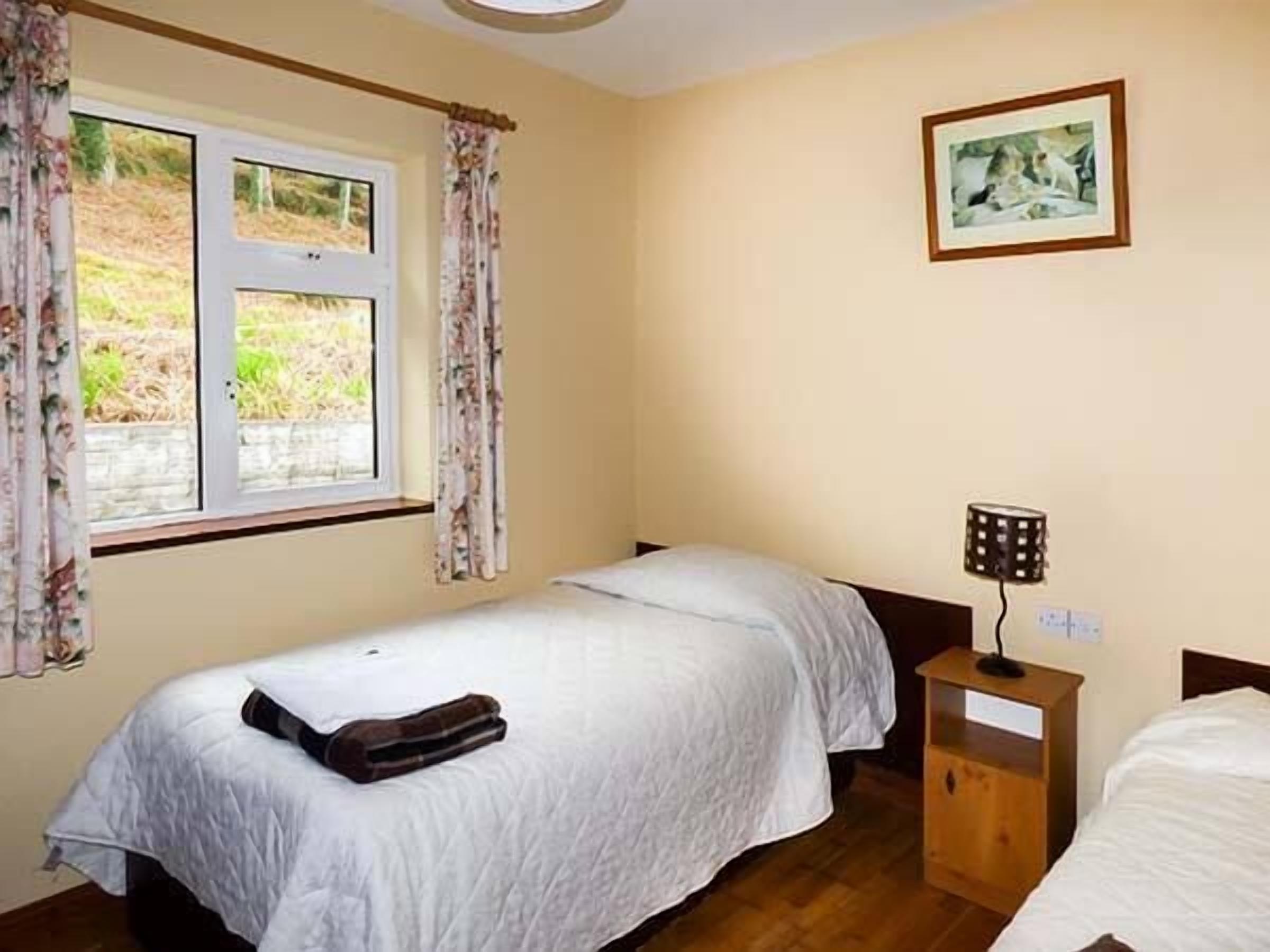 A bedroom with two single beds, a wooden nightstand with a lamp, a window with curtains, and a framed picture on the wall.