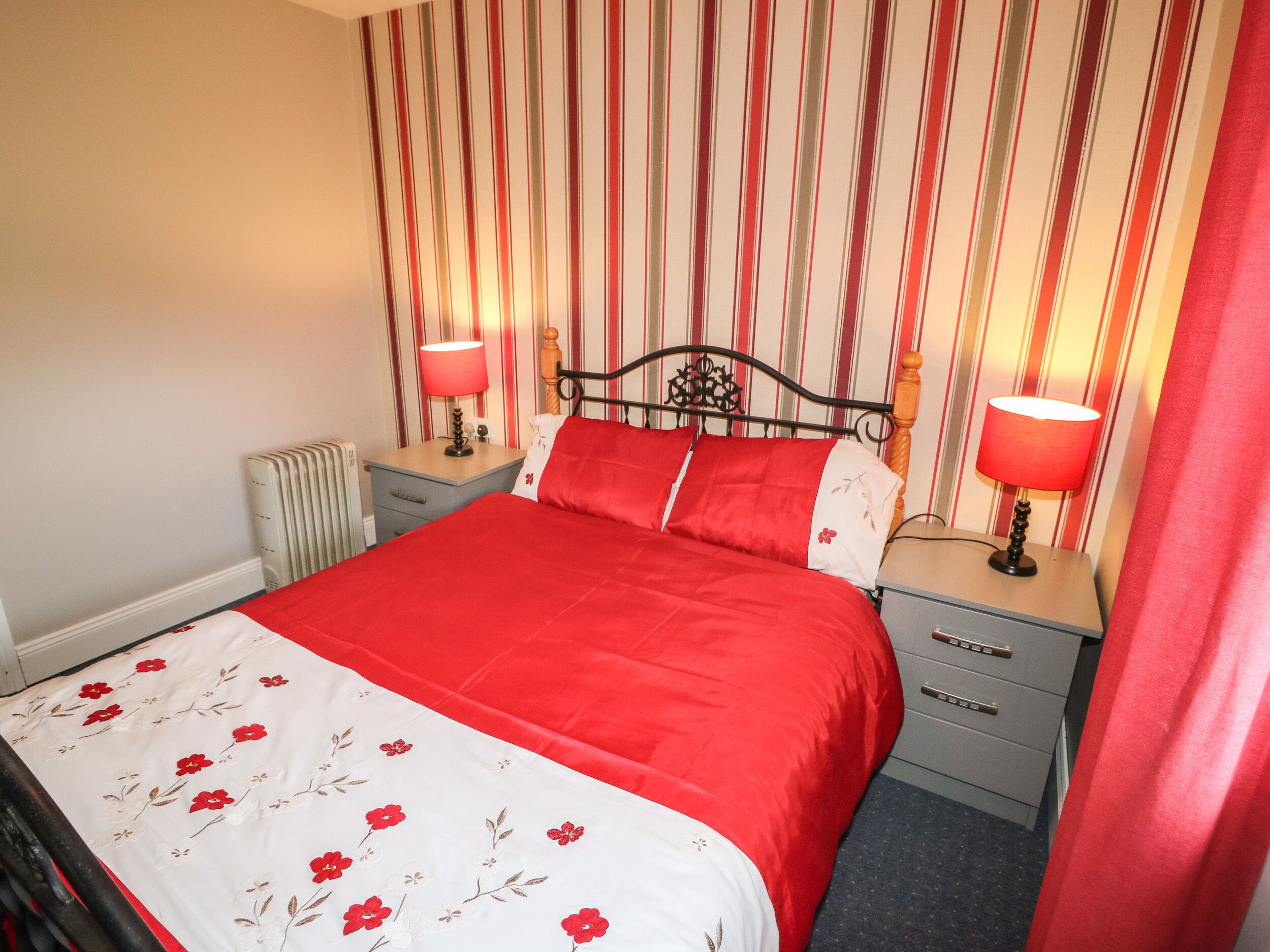 Ein ordentlich bezogenes Bett mit einer rot-weißen Blumenbettdecke, zwei Nachttischlampen, einem Holzkopfteil und einem gestreiften Wandhintergrund.