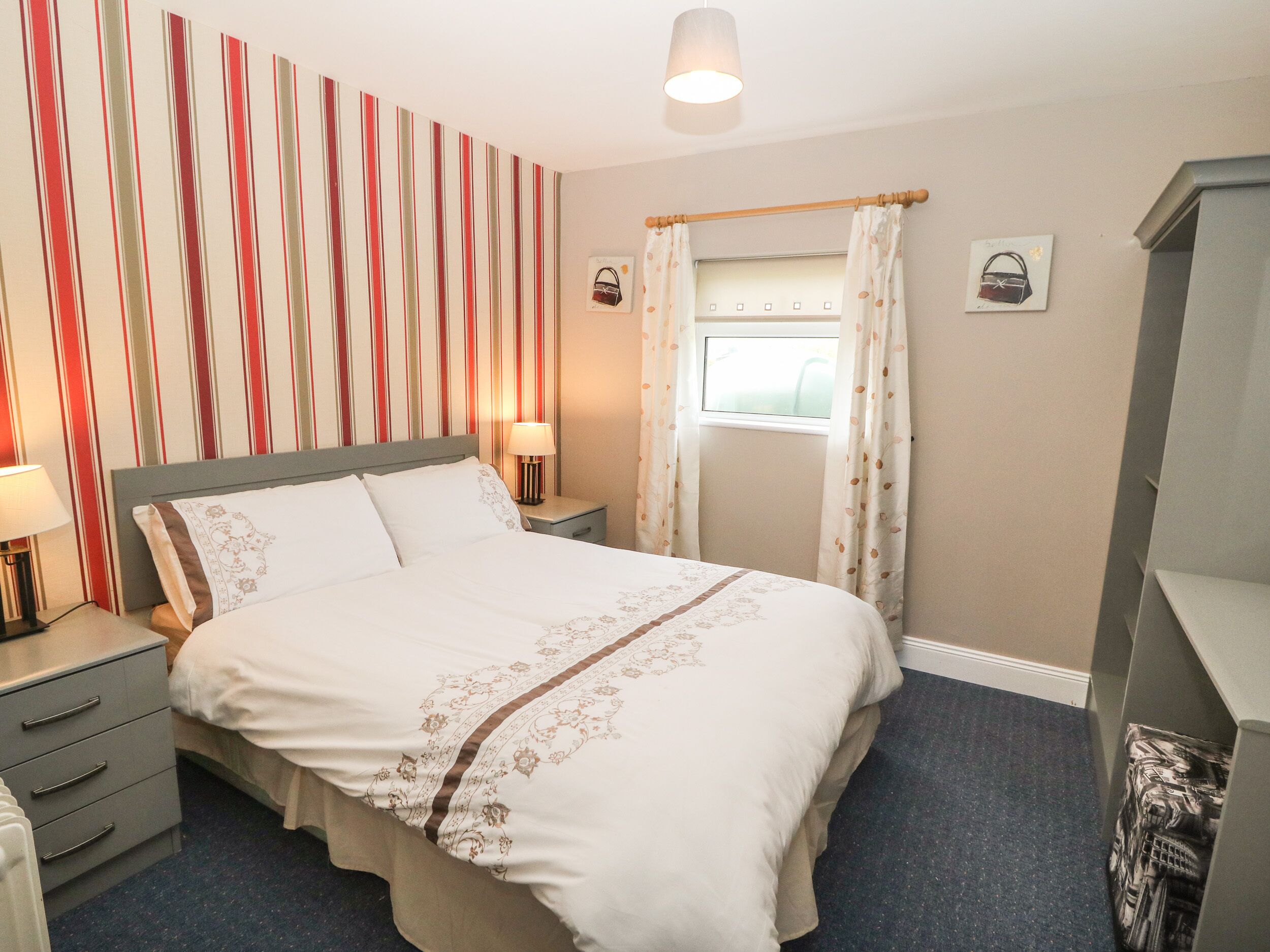 A neatly arranged hotel room with a bed, bedside tables, a lamp, a window with curtains, and a striped wall.
