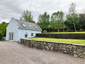 Une maison bleu clair avec un mur en pierre, une allée en gravier et une zone herbeuse avec des arbres.
