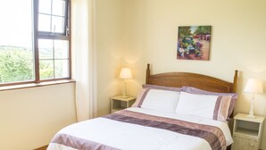 A neatly made bed with a wooden headboard, two bedside tables with lamps, a window with curtains, and a painting on the wall. - Flynn's Cottage (Miltown Malbay)