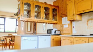 A kitchen with wooden cabinets, a microwave, a refrigerator, and a dining area with chairs. - Flynn's Cottage (Miltown Malbay)