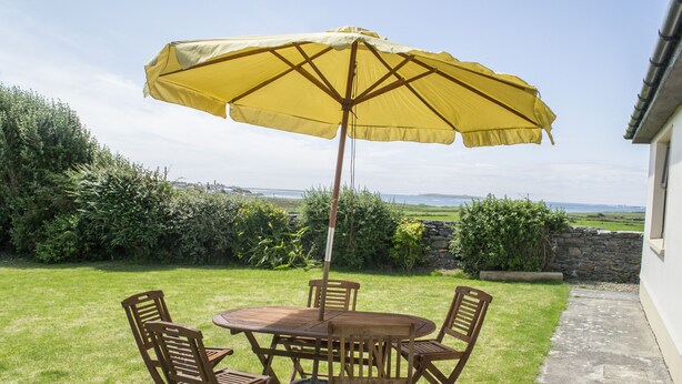 A patio with a round wooden table and four chairs, an umbrella, and a view of the sea and hills. - Flynn's Cottage (Miltown Malbay)