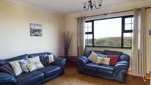A living room with two dark blue leather sofas, a wooden floor, a chandelier, a window with curtains, and a picture on the wall. - Flynn's Cottage (Miltown Malbay)