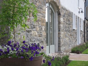 A stone building with a large planter of purple flowers in front. - Old Village Barn (Woodford)