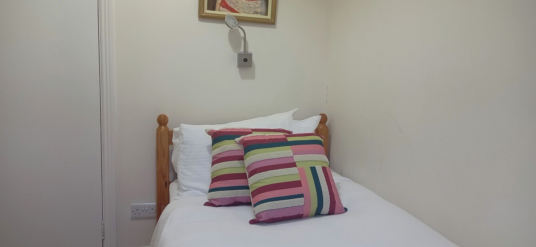 A small, single-bed room with a wooden headboard, white bedding, and colorful striped pillows.