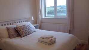 A neatly made bed with pillows and folded towels, a bedside lamp, and a view of a coastal landscape through a window.