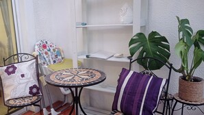 A small patio area with a round table, two chairs, a shelf with decorative items, and a large plant.