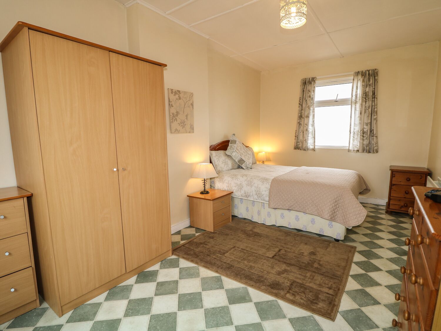A bedroom with a wooden wardrobe, a bed with a floral bedspread, a nightstand with a lamp, a window with curtains, and a checkered floor.