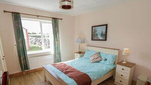 A bedroom with a bed, a nightstand, a lamp, a window with curtains, a radiator, and a painting on the wall. - Lough Mask Road Fishing Lodge (Cong)