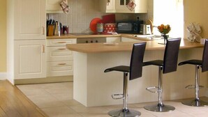 Una cocina con armarios blancos, microondas y cafetera. Hay tres taburetes de bar y una ventana con cortinas.