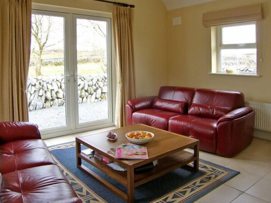 A living room with a red leather sofa, a wooden coffee table with fruits and magazines, a patterned rug, and a view of a stone wall and trees through a glass door.