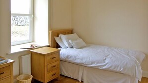 Un dormitorio con una cama individual, una mesita de noche de madera, una mesilla auxiliar y una ventana con vistas a un muro de piedra.