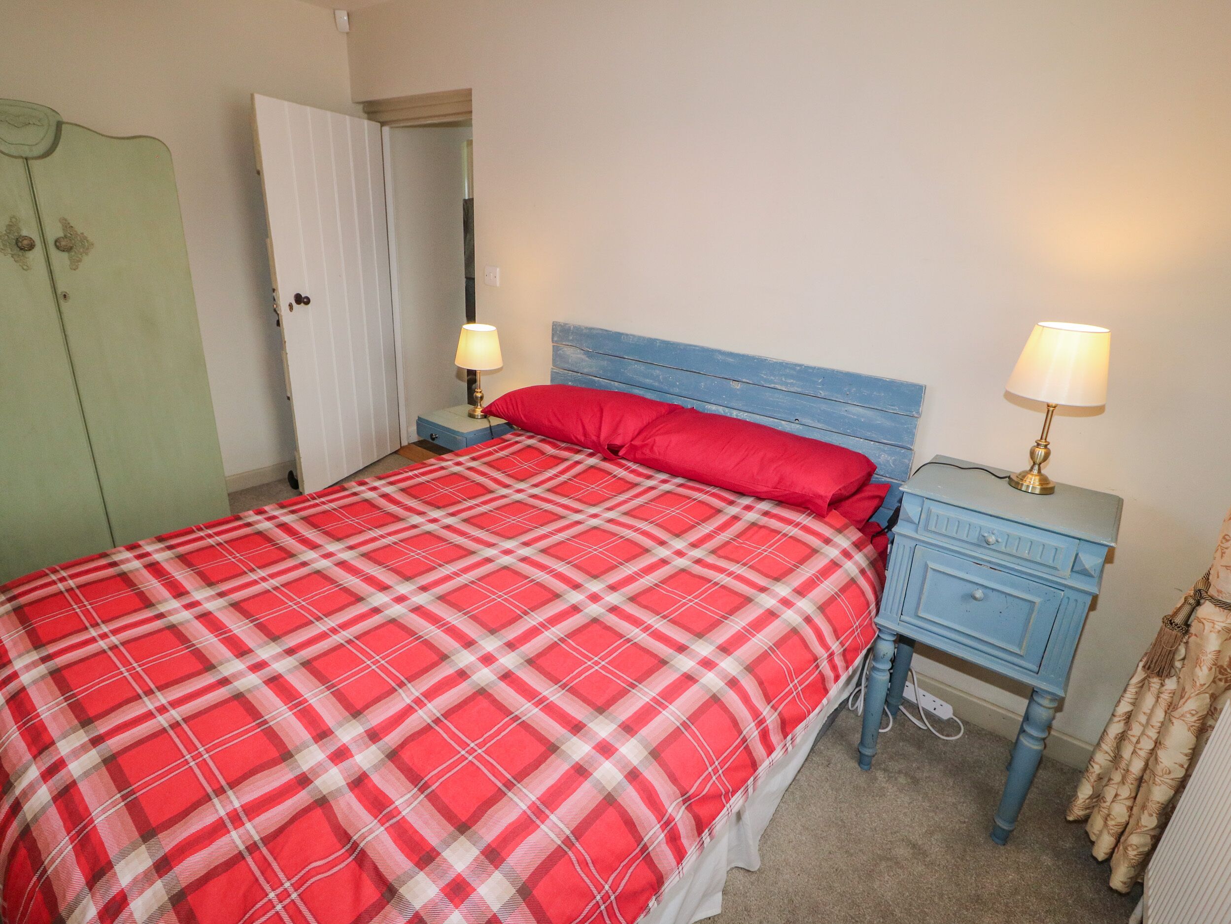 A bedroom with a red plaid bedspread, a blue headboard, two bedside tables with lamps, and a green wardrobe.