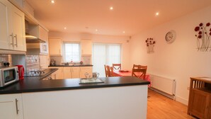 A modern kitchen with white cabinets, a black countertop, and a red kettle. There is a dining area with a red and white checkered tablecloth and wooden chairs. A clock and a vase with flowers are on the wall. - 63 Moinin (Kilkee)