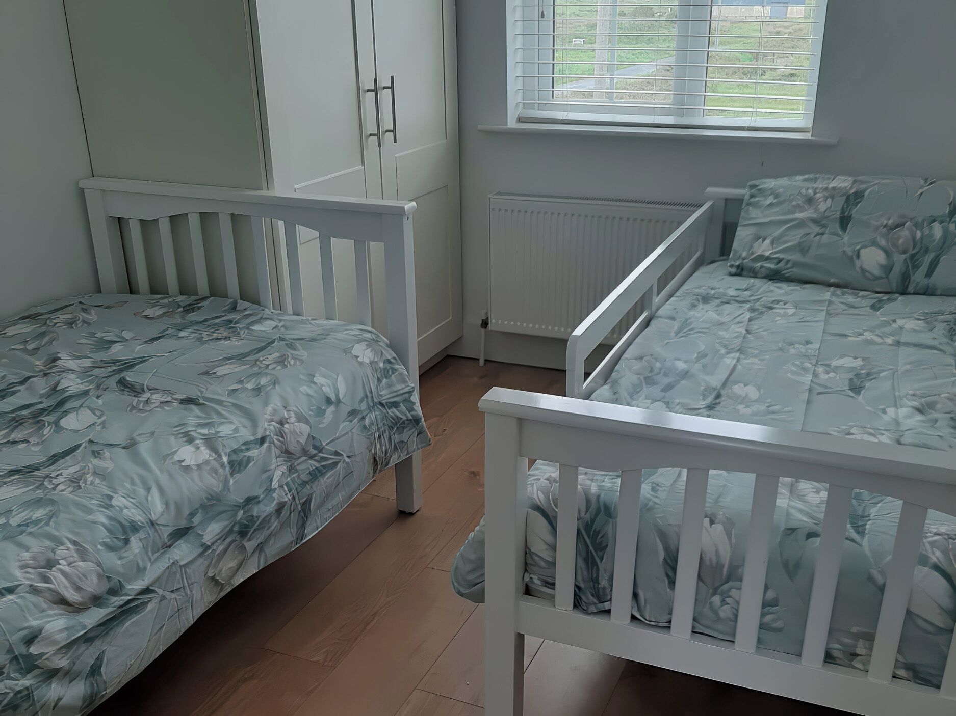 A bedroom with two single beds, a wardrobe, a radiator, and a window with blinds.