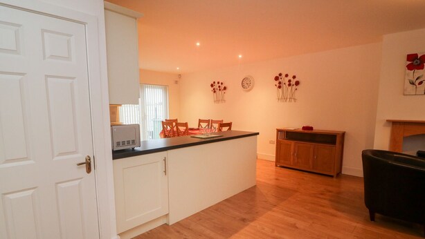 A kitchen with white cabinets, a black countertop, and wooden flooring. There is a dining area with chairs and a table, and a clock on the wall. - 63 Moinin (Kilkee)