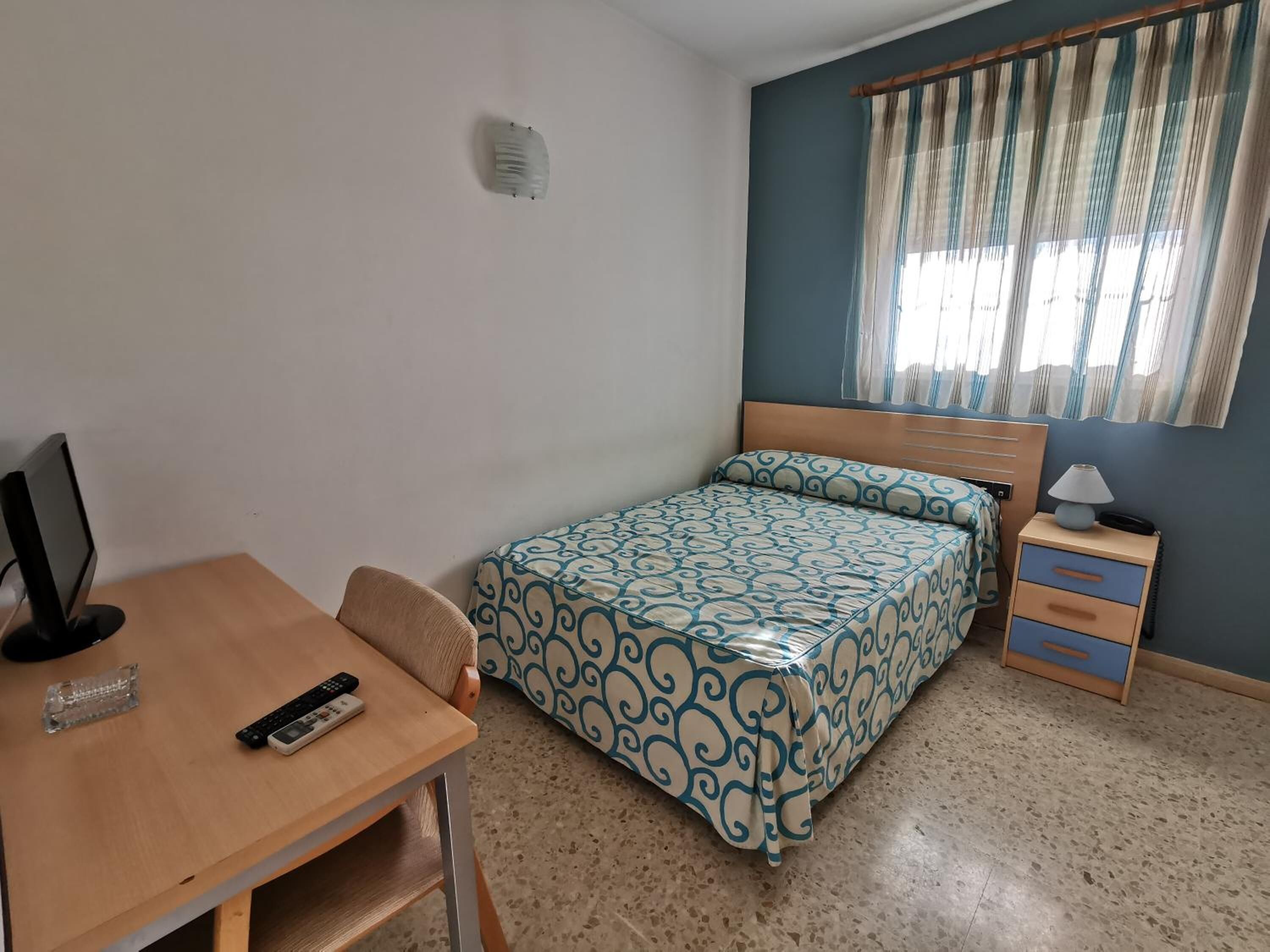 Un dormitorio con cama, escritorio con monitor de computadora y controles remotos, una silla, mesita de noche con lámpara y ventana con cortinas.