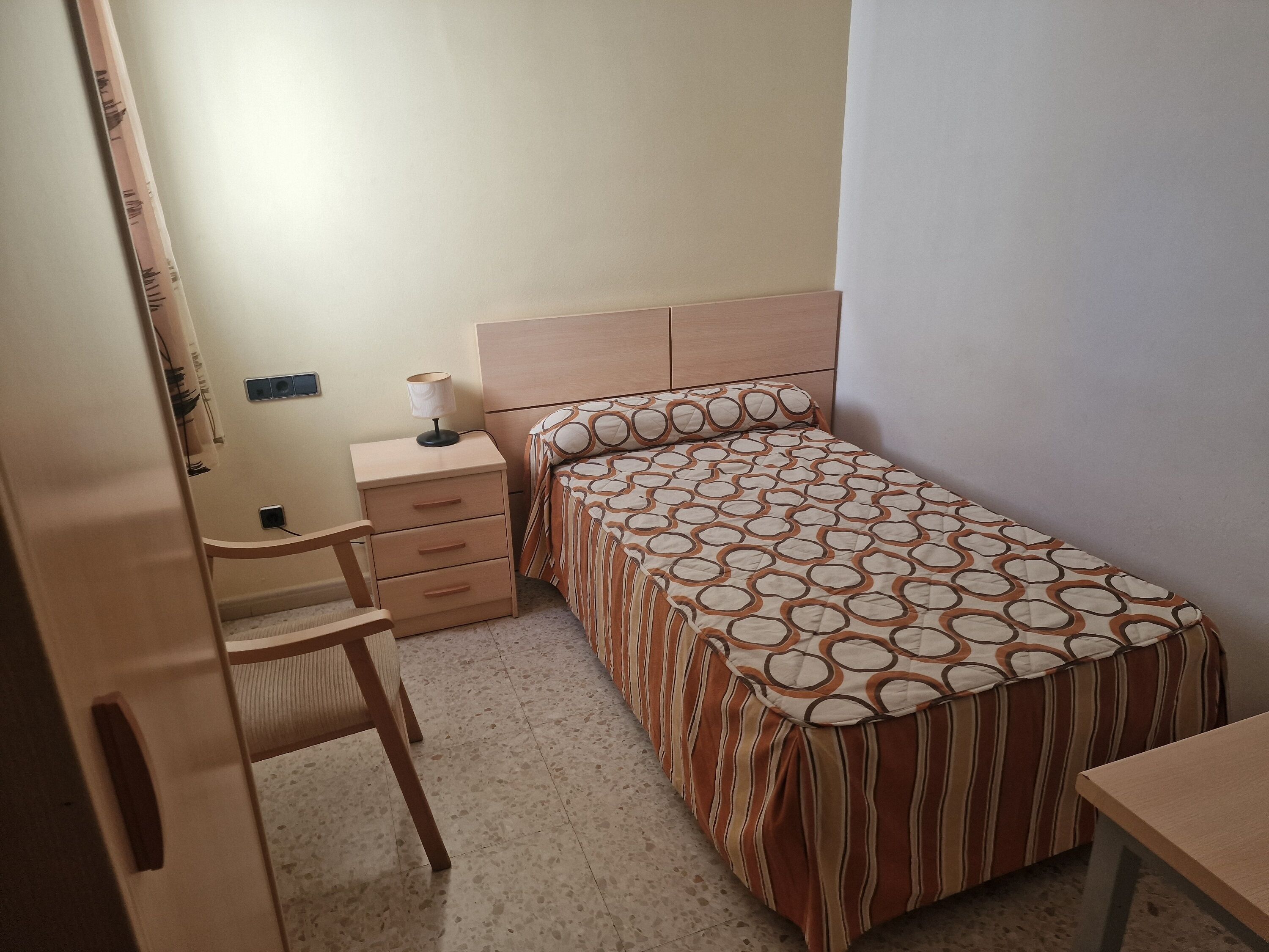 Een kleine, verzorgde slaapkamer met een enkel bed, een nachtkastje met een lamp, een stoel en een garderobe.